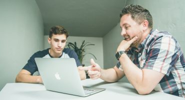 man wearing white and black plaid button-up sports shirt pointing the silver MacBook