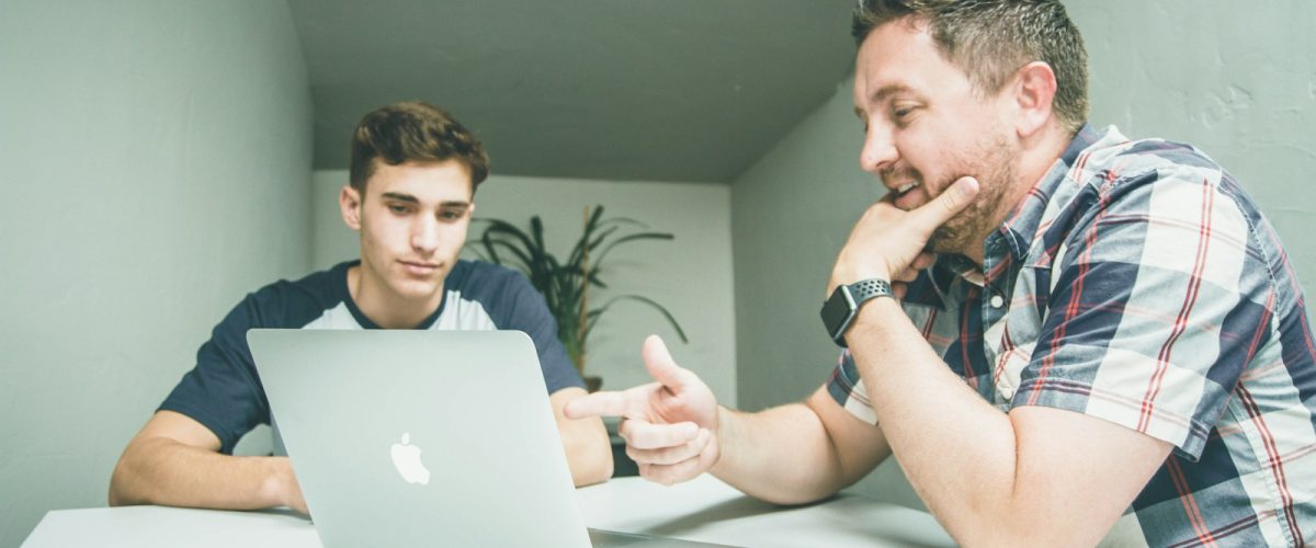 man wearing white and black plaid button-up sports shirt pointing the silver MacBook